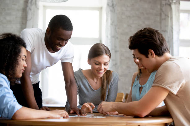 How to Make Friends as an Adult. Happy multiracial friends assembling puzzle jigsaw at home or in cafe, teamwork concept, diverse laughing students sitting at table, spending weekend together, weekend activities, having fun.